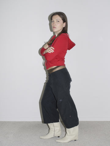 Model wearing a fitted red zip-up hoodie with studded hood detail, low-rise black Nike cargo pants, and pointed white leather cowboy boots. The look captures peak Y2K attitude — part Britney, part G-Star, part Western revival.