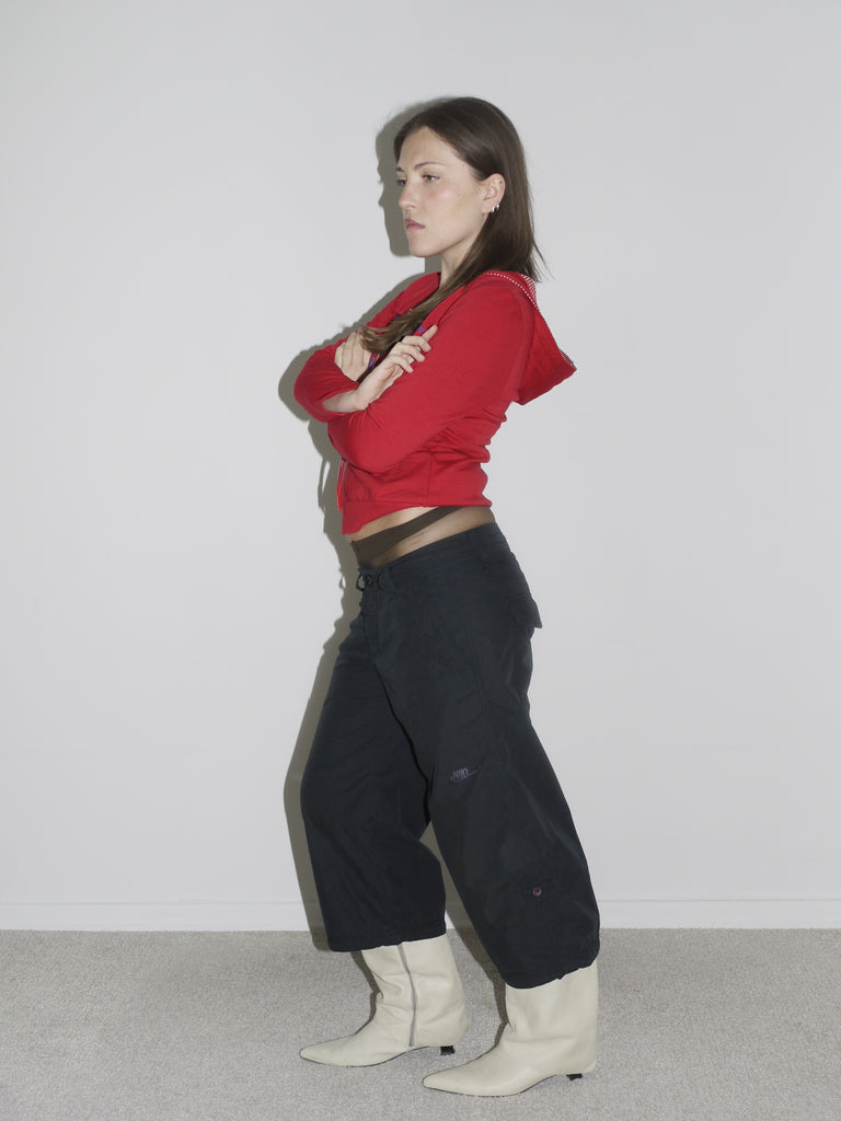 Model wearing a fitted red zip-up hoodie with studded hood detail, low-rise black Nike cargo pants, and pointed white leather cowboy boots. The look captures peak Y2K attitude — part Britney, part G-Star, part Western revival.