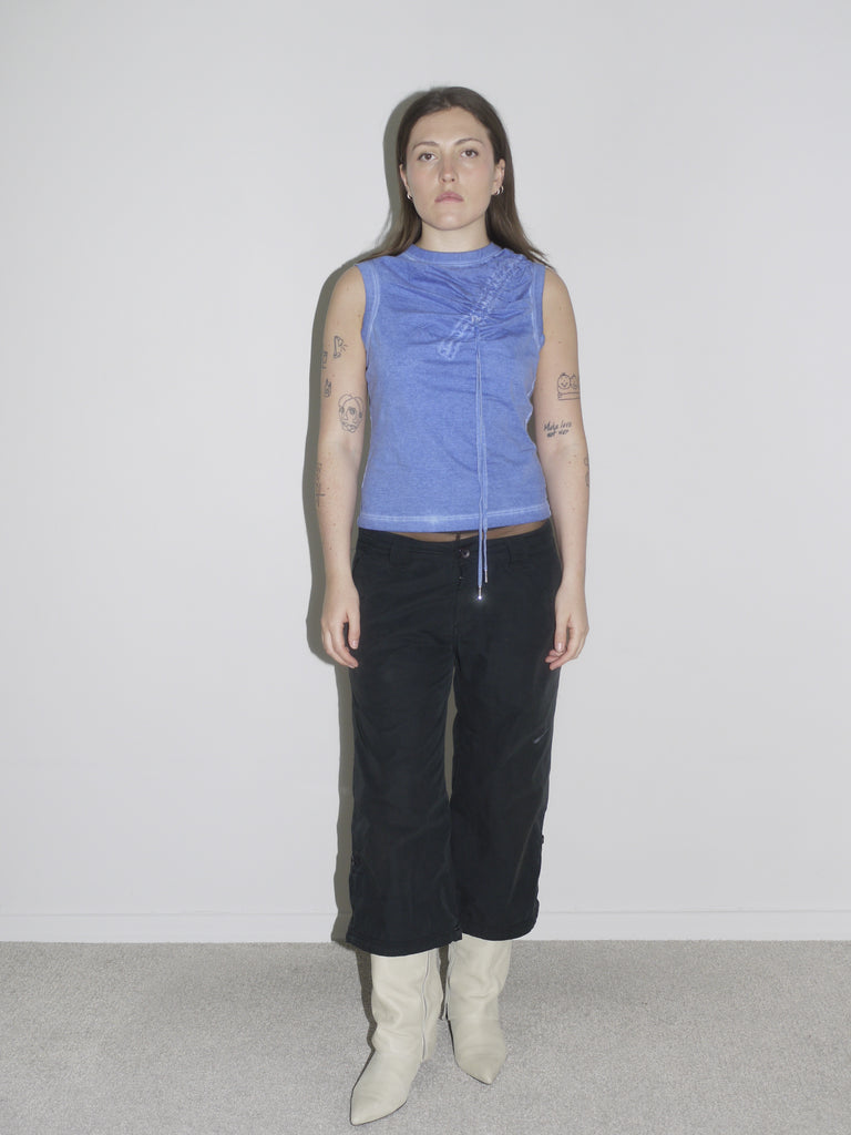 Model wearing a fitted blue sleeveless top with gathered drawstring detail, low-rise black cargo pants, and pointed white leather cowboy boots. The look captures early 2000s sexiness and attitude — a strong nod to vintage Dsquared2, with Western and utilitarian crossover
