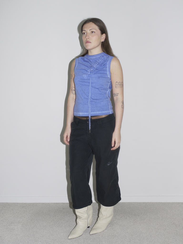 Model wearing a fitted blue sleeveless top with gathered drawstring detail, low-rise black cargo pants, and pointed white leather cowboy boots. The look captures early 2000s sexiness and attitude — a strong nod to vintage Dsquared2, with Western and utilitarian crossover