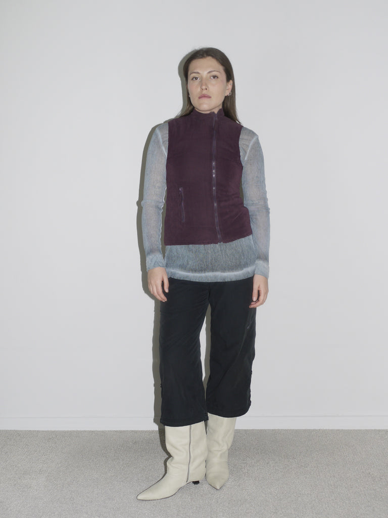 Model wearing a sheer blue long-sleeve top layered under a deep plum fleece zip-up vest, paired with black cropped cargo pants and pointed white leather boots. A soft-meets-technical combo, echoing early 2000s layering aesthetics — think Diesel, MM6, and vintage mountaineering gear.