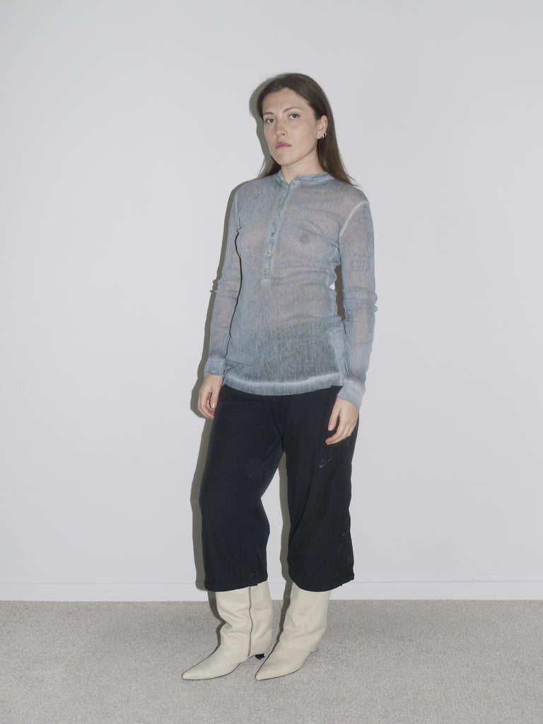 Model wearing a sheer ice-blue long-sleeve henley top with front button detail, styled with low-rise black cargo pants and pointed white cowboy boots. The look blends fragility with function — early 2000s Helmut Lang vibes meet softcore transparency.