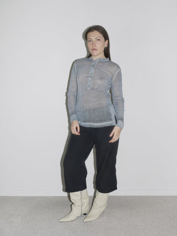 Model wearing a sheer ice-blue long-sleeve henley top with front button detail, styled with low-rise black cargo pants and pointed white cowboy boots. The look blends fragility with function — early 2000s Helmut Lang vibes meet softcore transparency.