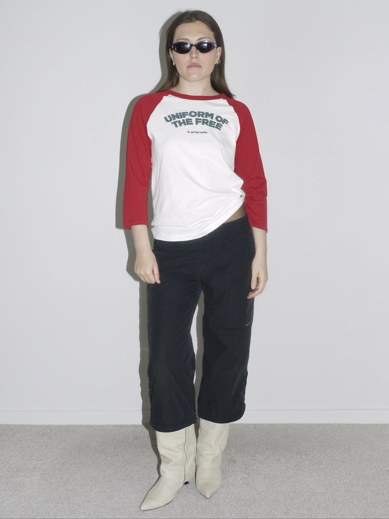 Model wearing a red and white raglan shirt with “Uniform of the Free” print by G-Star RAW, paired with black cropped cargo pants and pointed white cowboy boots. Styled with oval black sunglasses — sporty, rebellious and straight out of a 2000s campaign.