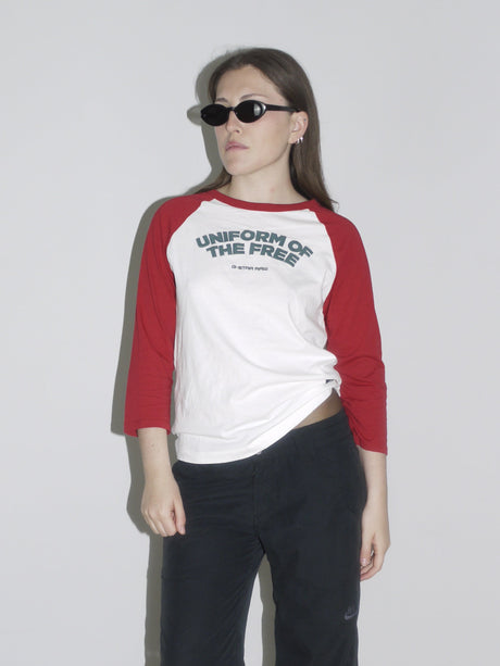 Model wearing a red and white raglan shirt with “Uniform of the Free” print by G-Star RAW, paired with black cropped cargo pants and pointed white cowboy boots. Styled with oval black sunglasses — sporty, rebellious and straight out of a 2000s campaign.