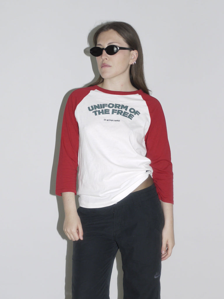 Model wearing a red and white raglan shirt with “Uniform of the Free” print by G-Star RAW, paired with black cropped cargo pants and pointed white cowboy boots. Styled with oval black sunglasses — sporty, rebellious and straight out of a 2000s campaign.