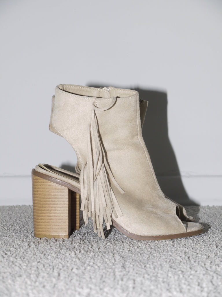Close-up of beige suede open-toe heels with chunky stacked wooden heel and dramatic fringe detail along the side. Western-inspired and 2000s glam — think Isabel Marant meets early Zara Studio with festival edge.