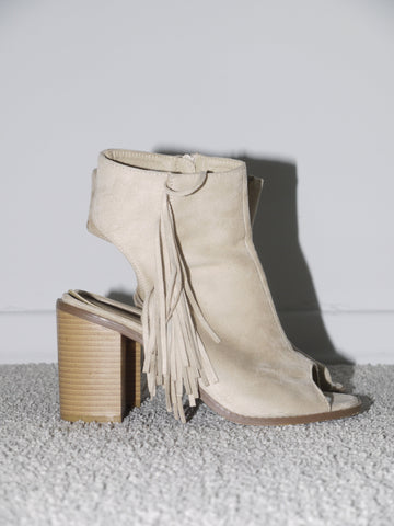 Close-up of beige suede open-toe heels with chunky stacked wooden heel and dramatic fringe detail along the side. Western-inspired and 2000s glam — think Isabel Marant meets early Zara Studio with festival edge.
