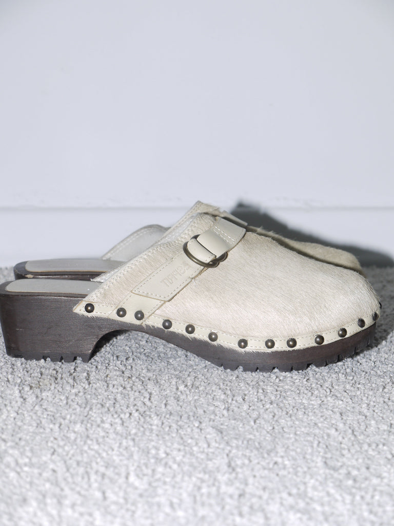 Close-up of beige wooden-sole clogs made of pony hair leather, featuring a metal buckle strap and antique-style stud detailing along the edge — vintage-inspired footwear blending rustic charm with 2000s minimalism, reminiscent of Marni and Maison Margiela.