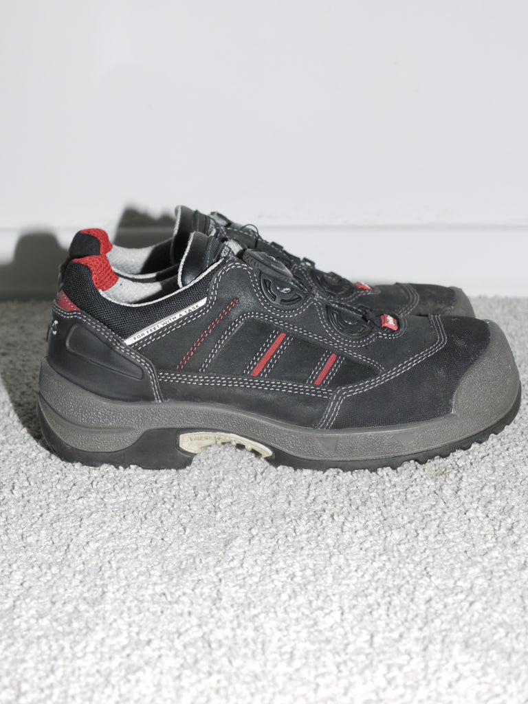 
Close-up of a pair of chunky black safety-style sneakers with red and grey detailing, thick sole, and reinforced toe — reminiscent of Salomon and early 2000s utility footwear. Industrial workwear meets techwear aesthetic.