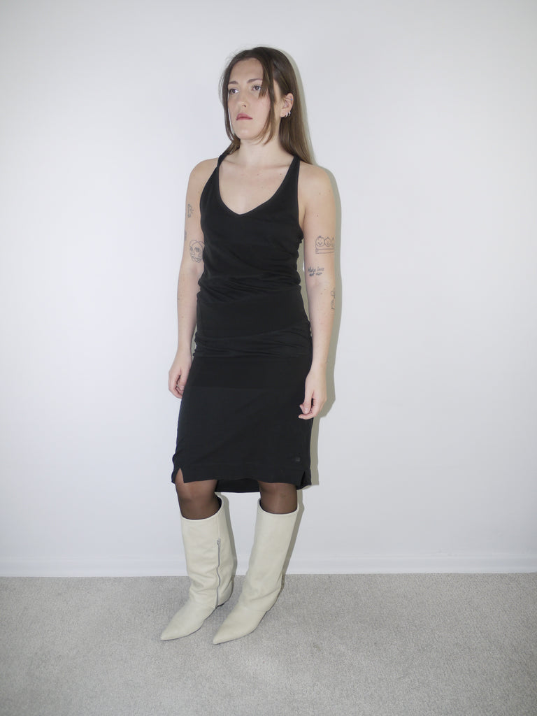 Model wearing a black fitted G-Star RAW tank dress with side logo tag and small front slit, styled with sheer black tights and cream pointed knee-high boots — minimal Y2K-inspired silhouette with subtle branding, shot against plain white background.