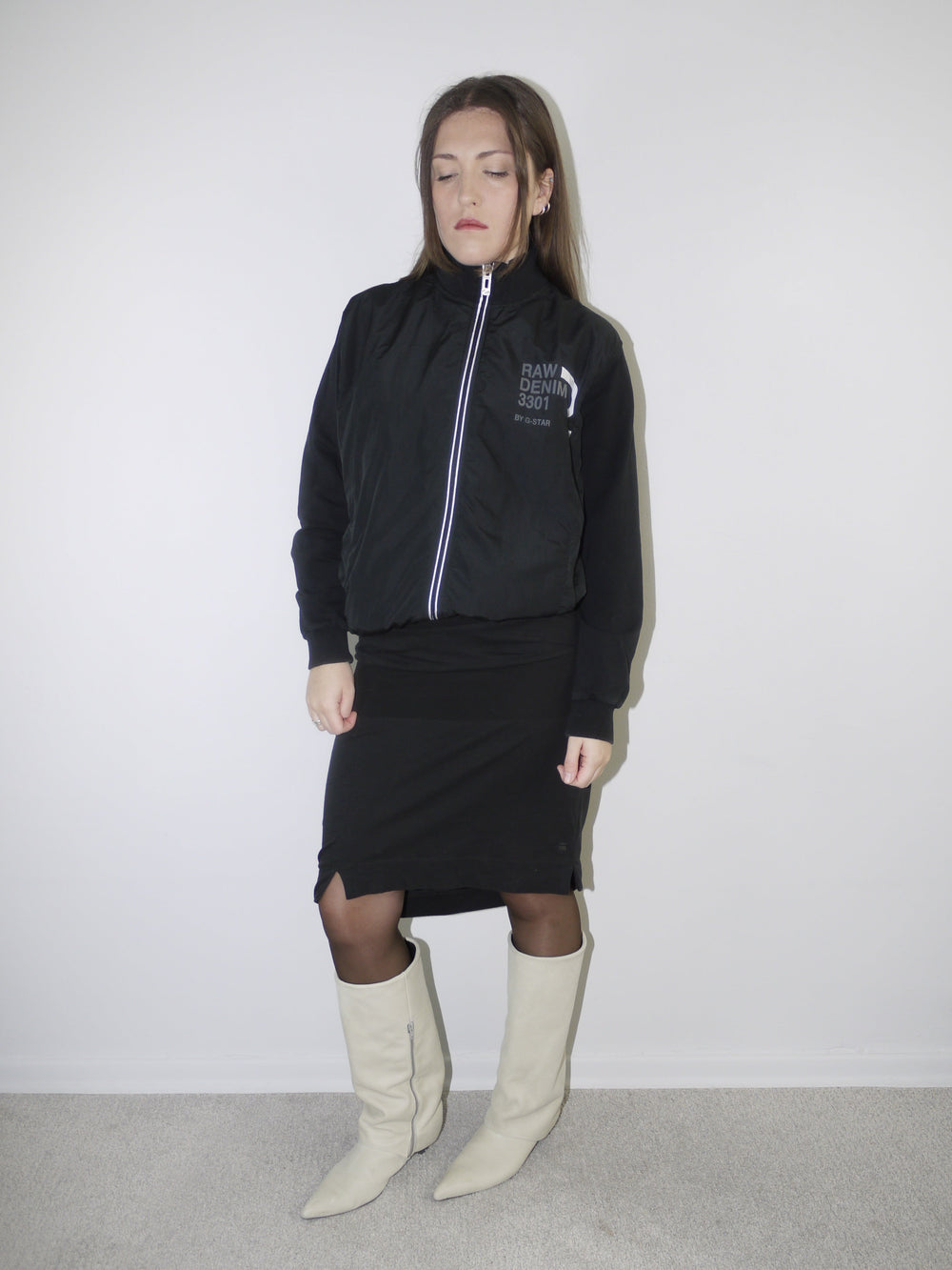 Model wearing a black G-Star RAW zip-up bomber jacket with logo print, paired with a fitted black midi skirt with front slit, sheer black tights and cream pointed knee-high boots — monochrome Y2K streetwear look with clean layering, photographed against white wall.