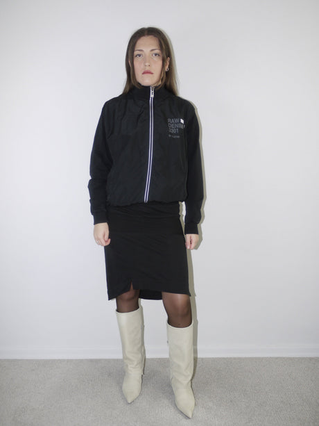 Model wearing a black G-Star RAW zip-up bomber jacket with logo print, paired with a fitted black midi skirt with front slit, sheer black tights and cream pointed knee-high boots — monochrome Y2K streetwear look with clean layering, photographed against white wall.