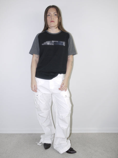 Model wearing a black and grey colorblock graphic T-shirt with printed text and wide white cargo pants, styled with pointed black flats — casual streetwear outfit with early 2000s attitude, photographed against white wall.

