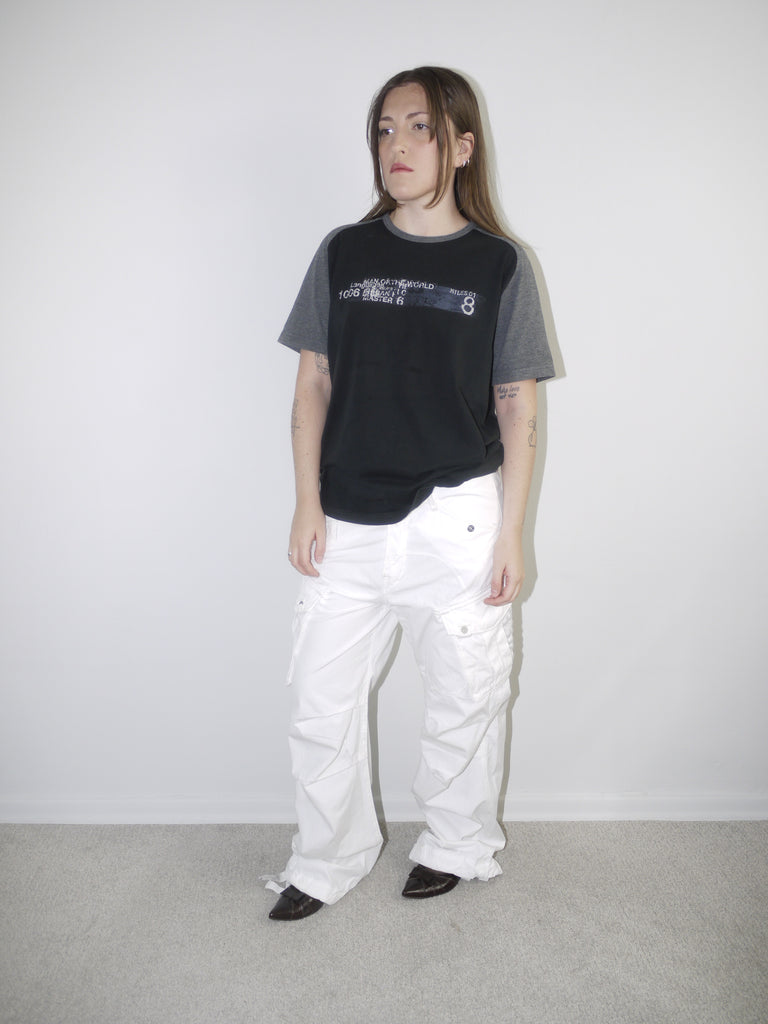 Model wearing a black and grey colorblock graphic T-shirt with printed text and wide white cargo pants, styled with pointed black flats — casual streetwear outfit with early 2000s attitude, photographed against white wall.

