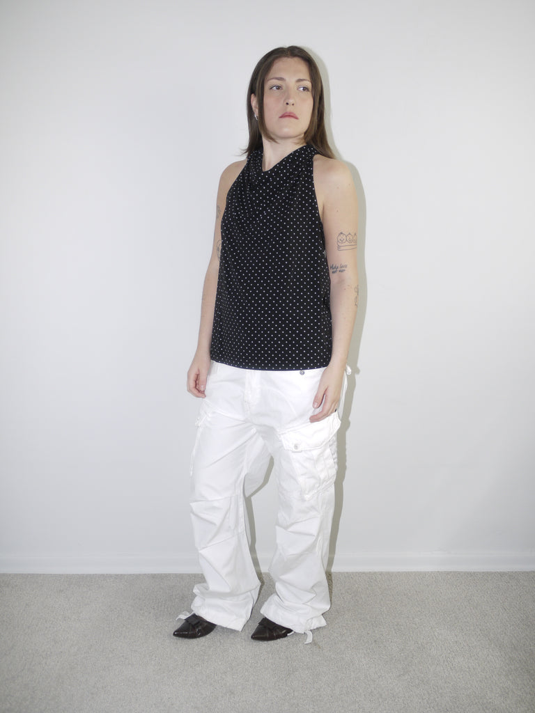 Model wearing a sleeveless black halterneck top with fine white polka dots, styled with white wide-leg cargo trousers and pointed flats — playful yet clean Y2K-inspired look, photographed against plain white background.