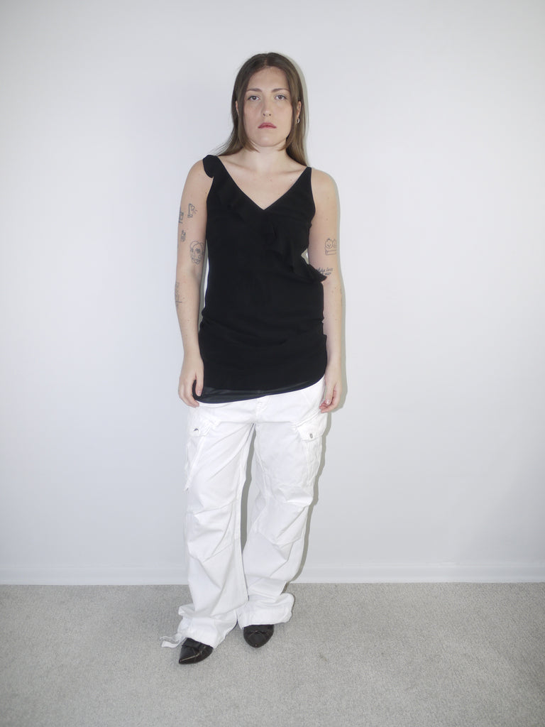 Model wearing a black asymmetrical waterfall draped top styled as a mini dress, paired with white utility cargo pants and pointed black heels — deconstructed Y2K-inspired layering look with soft volume and streetwear edge, shot against plain white wall.
