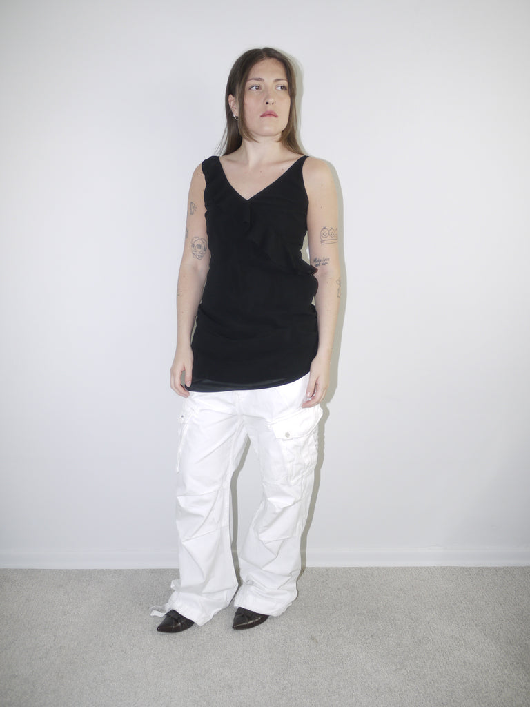 Model wearing a black asymmetrical waterfall draped top styled as a mini dress, paired with white utility cargo pants and pointed black heels — deconstructed Y2K-inspired layering look with soft volume and streetwear edge, shot against plain white wall.