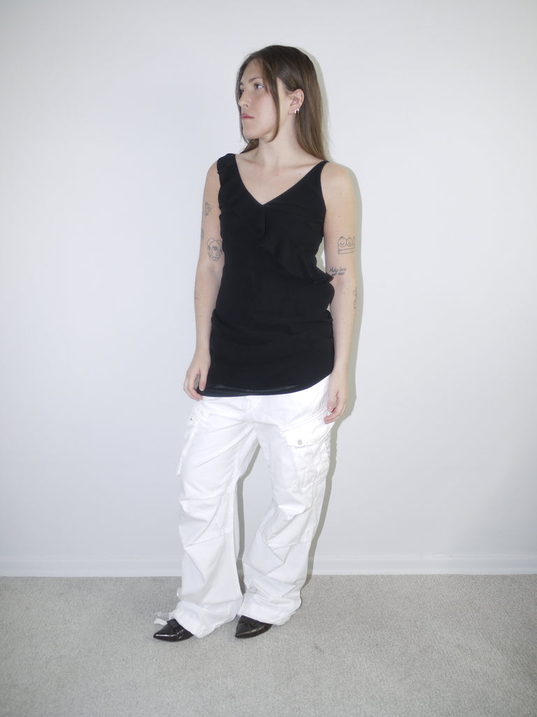 Model wearing a black asymmetrical waterfall draped top styled as a mini dress, paired with white utility cargo pants and pointed black heels — deconstructed Y2K-inspired layering look with soft volume and streetwear edge, shot against plain white wall.