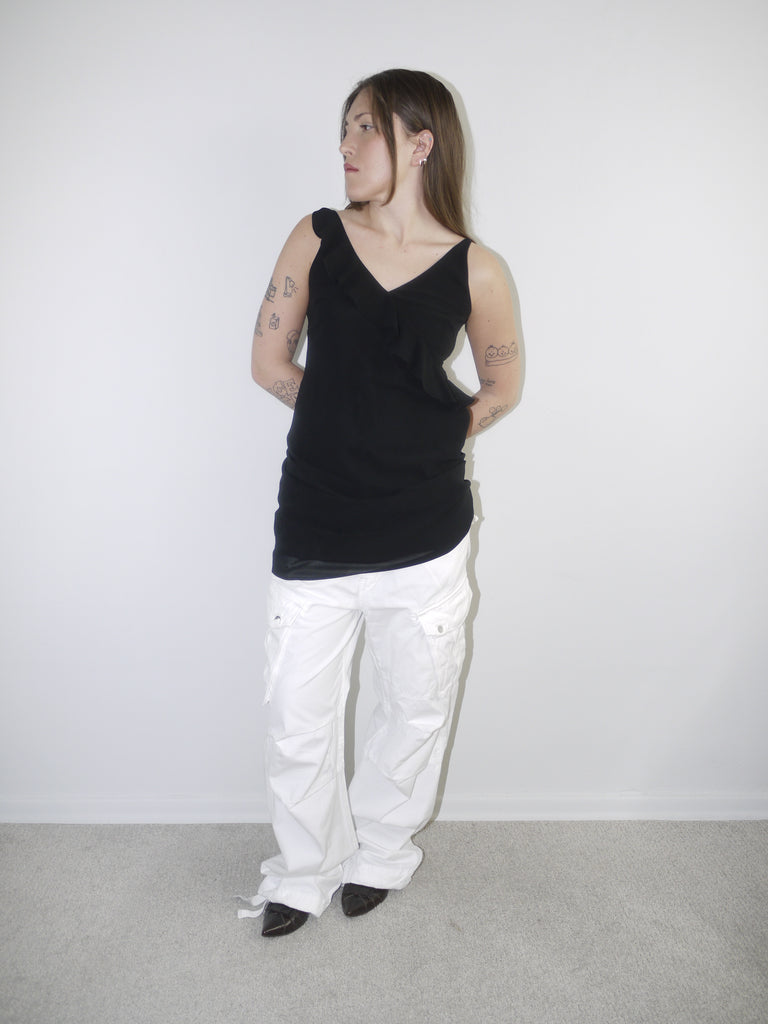 Model wearing a black asymmetrical waterfall draped top styled as a mini dress, paired with white utility cargo pants and pointed black heels — deconstructed Y2K-inspired layering look with soft volume and streetwear edge, shot against plain white wall.