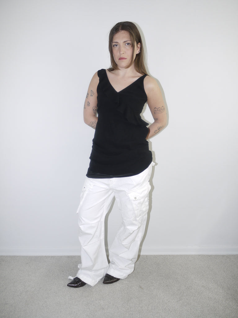 Model wearing a black asymmetrical waterfall draped top styled as a mini dress, paired with white utility cargo pants and pointed black heels — deconstructed Y2K-inspired layering look with soft volume and streetwear edge, shot against plain white wall.