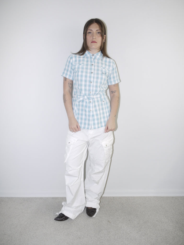 Model wearing a vintage Jack Wolfskin short-sleeve checkered button-up shirt in light blue and white, cinched at the waist with tie detail, styled with white wide-leg cargo trousers and pointed black heels — functional meets feminine in a Y2K-inspired outdoor look, photographed against clean white wall.