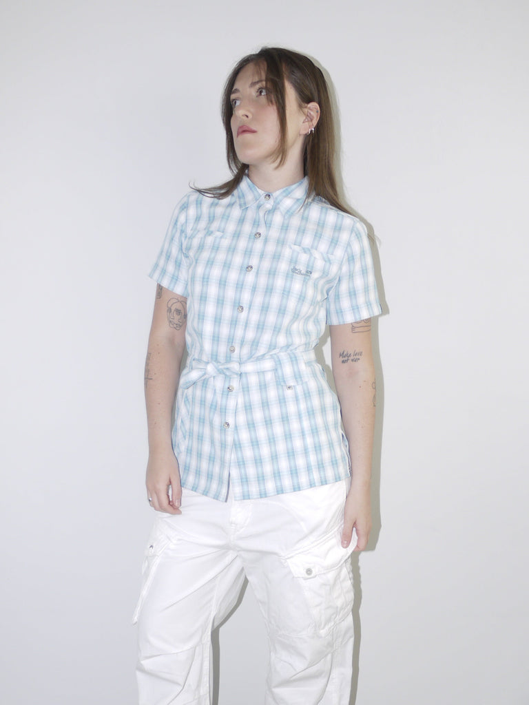 Model wearing a vintage Jack Wolfskin short-sleeve checkered button-up shirt in light blue and white, cinched at the waist with tie detail, styled with white wide-leg cargo trousers and pointed black heels — functional meets feminine in a Y2K-inspired outdoor look, photographed against clean white wall.