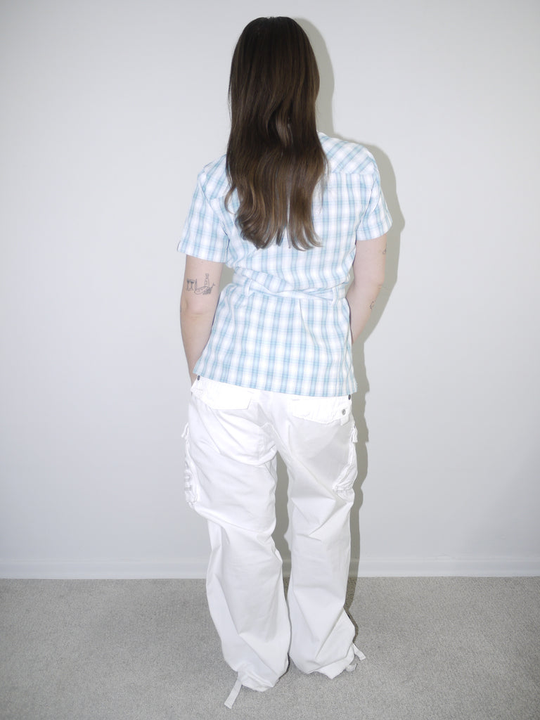 Model wearing a vintage Jack Wolfskin short-sleeve checkered button-up shirt in light blue and white, cinched at the waist with tie detail, styled with white wide-leg cargo trousers and pointed black heels — functional meets feminine in a Y2K-inspired outdoor look, photographed against clean white wall.