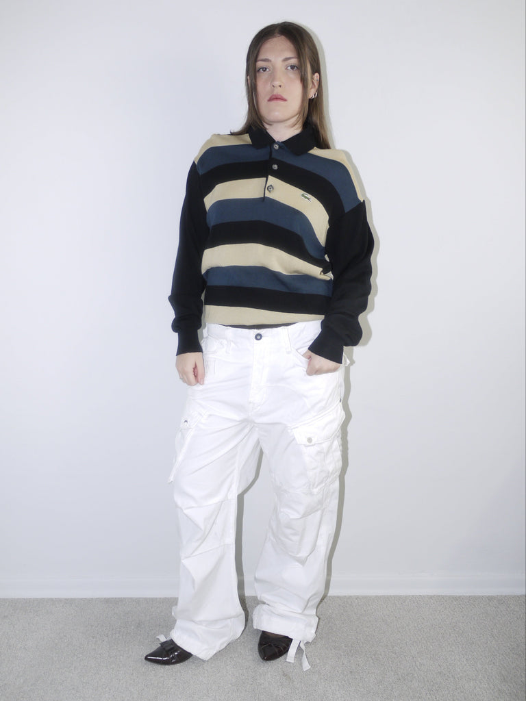 Model wearing a vintage Lacoste striped knit polo sweater in navy, beige and black, paired with wide-leg white drawstring cargo pants and pointed black heels — relaxed retro sportswear look with early 2000s energy, shot against neutral background.