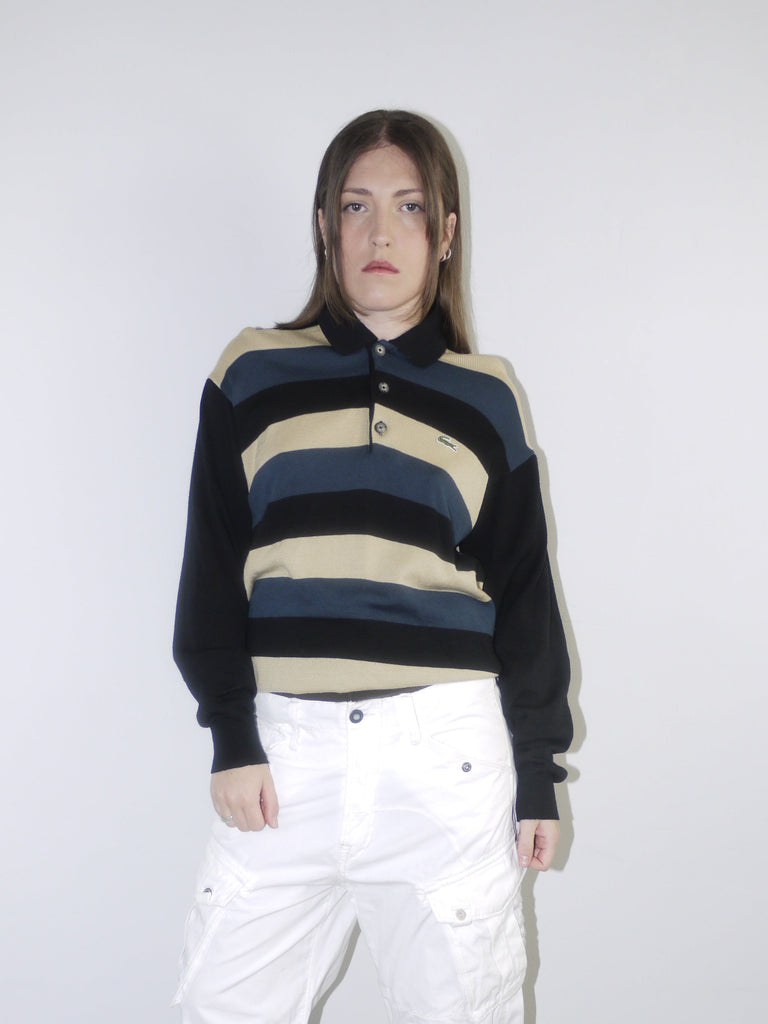 Model wearing a vintage Lacoste striped knit polo sweater in navy, beige and black, paired with wide-leg white drawstring cargo pants and pointed black heels — relaxed retro sportswear look with early 2000s energy, shot against neutral background.