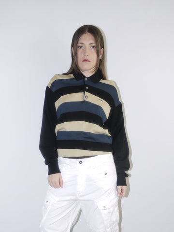 Model wearing a vintage Lacoste striped knit polo sweater in navy, beige and black, paired with wide-leg white drawstring cargo pants and pointed black heels — relaxed retro sportswear look with early 2000s energy, shot against neutral background.