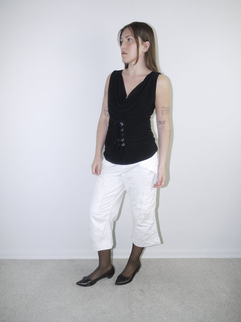 Model wearing a black sleeveless corset-style top with draped neckline and lace-up waist detail, styled with cropped white cargo pants, sheer black tights and pointed patent flats — bold Y2K-inspired silhouette with minimal styling, photographed against white background.
