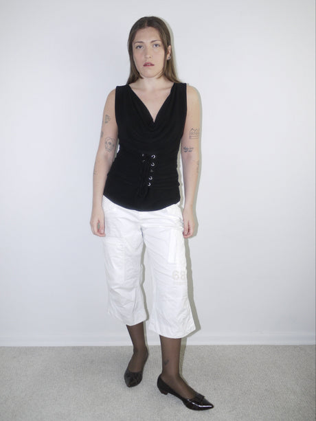 Model wearing a black sleeveless corset-style top with draped neckline and lace-up waist detail, styled with cropped white cargo pants, sheer black tights and pointed patent flats — bold Y2K-inspired silhouette with minimal styling, photographed against white background.