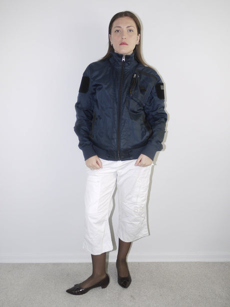 White cropped cargo pants styled with sheer black tights, dark patent leather pointed flats and an oversized periwinkle blue knit sweater — modern casual outfit with Y2K elements, photographed against neutral wall.