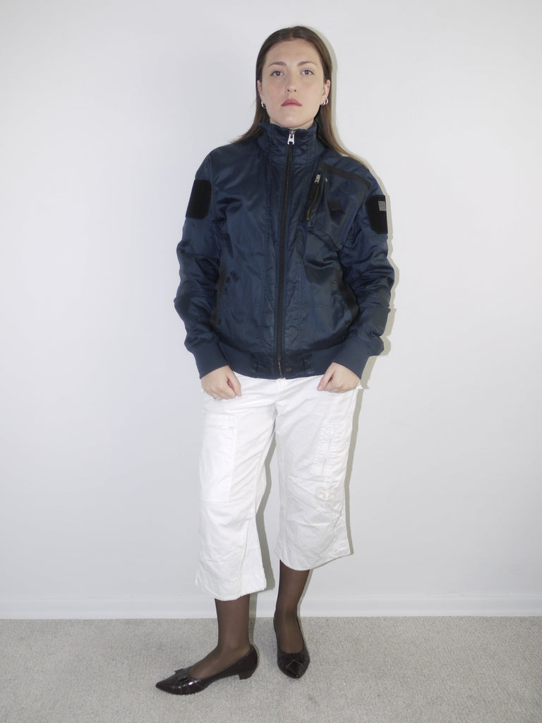 White cropped cargo pants styled with sheer black tights, dark patent leather pointed flats and an oversized periwinkle blue knit sweater — modern casual outfit with Y2K elements, photographed against neutral wall.