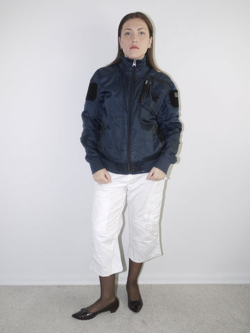 White cropped cargo pants styled with sheer black tights, dark patent leather pointed flats and an oversized periwinkle blue knit sweater — modern casual outfit with Y2K elements, photographed against neutral wall.