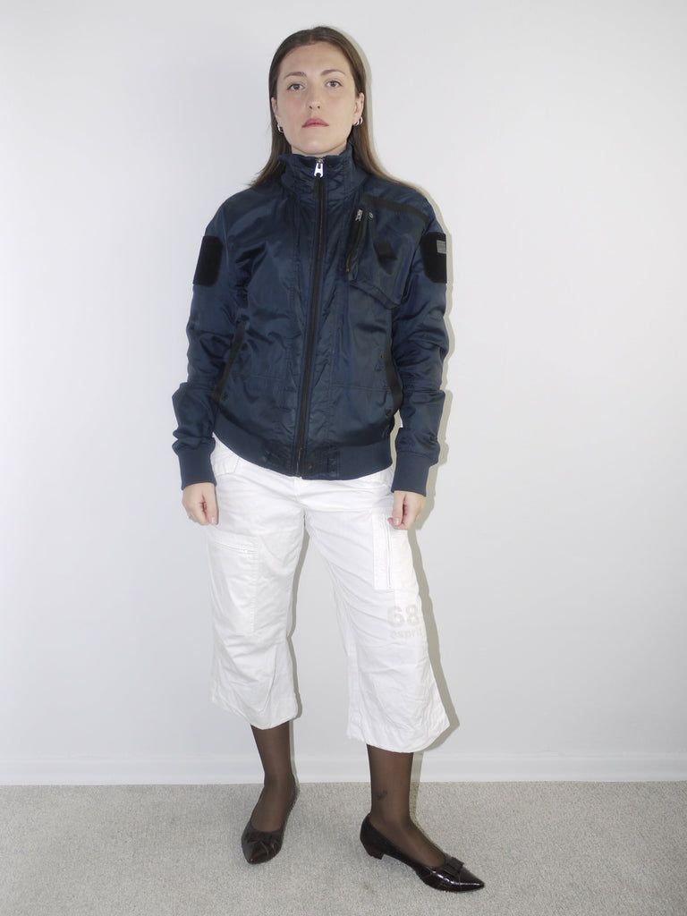 Model wearing a navy blue utility bomber jacket with high collar, paired with cropped white cargo pants, black tights and patent pointed flats — clean and edgy streetwear look with Y2K influence, shot against white backdrop.