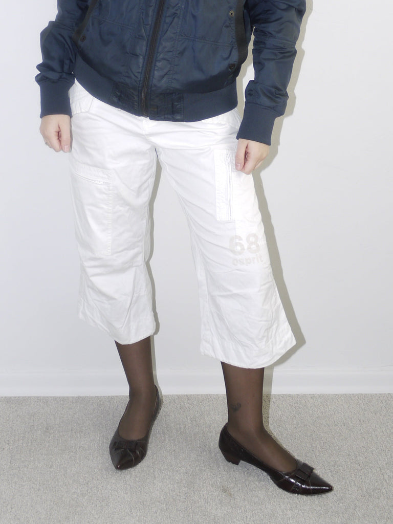 Model wearing a navy blue utility bomber jacket with high collar, paired with cropped white cargo pants, black tights and patent pointed flats — clean and edgy streetwear look with Y2K influence, shot against white backdrop.