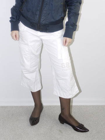 Model wearing a navy blue utility bomber jacket with high collar, paired with cropped white cargo pants, black tights and patent pointed flats — clean and edgy streetwear look with Y2K influence, shot against white backdrop.