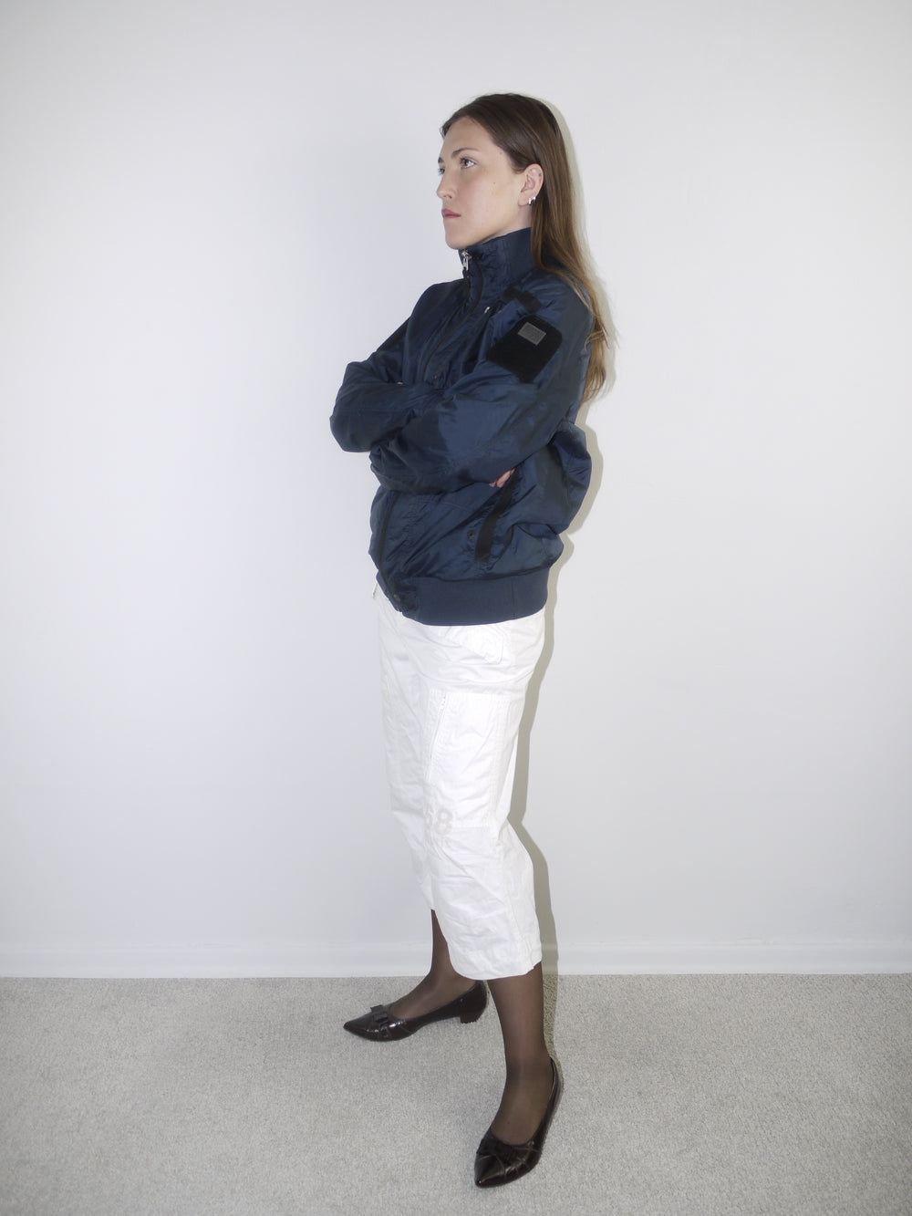 White cropped cargo pants styled with sheer black tights, dark patent leather pointed flats and an oversized periwinkle blue knit sweater — modern casual outfit with Y2K elements, photographed against neutral wall.