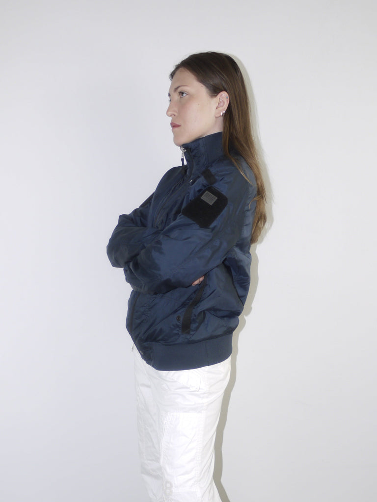 Model wearing a navy blue utility bomber jacket with high collar, paired with cropped white cargo pants, black tights and patent pointed flats — clean and edgy streetwear look with Y2K influence, shot against white backdrop.