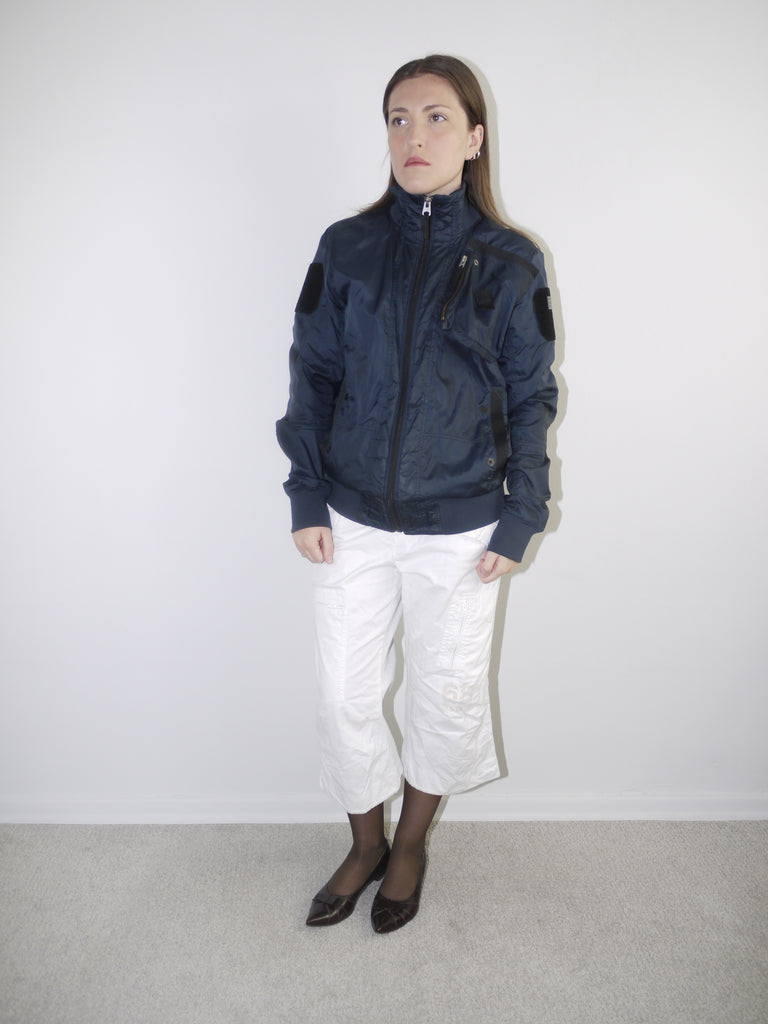 Model wearing a navy blue utility bomber jacket with high collar, paired with cropped white cargo pants, black tights and patent pointed flats — clean and edgy streetwear look with Y2K influence, shot against white backdrop.