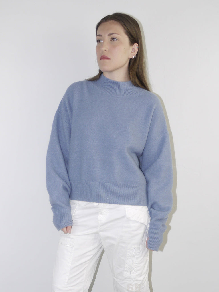 Model wearing a relaxed periwinkle blue knit sweater with structured shoulders, paired with white utility-style cropped pants — clean minimal outfit with soft color palette, shot against a neutral white background.