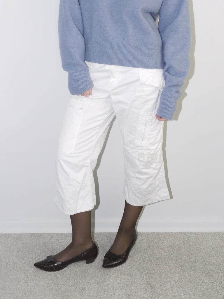 White cropped cargo pants styled with sheer black tights, dark patent leather pointed flats and an oversized periwinkle blue knit sweater — modern casual outfit with Y2K elements, photographed against neutral wall.