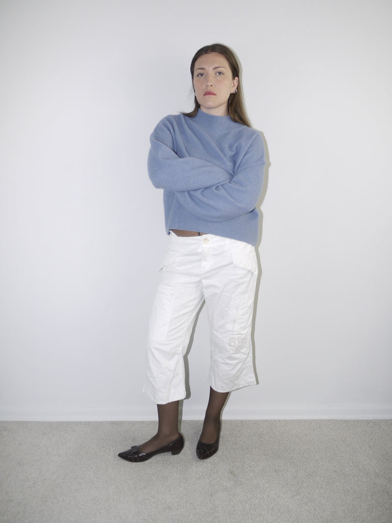 Model wearing a relaxed periwinkle blue knit sweater with structured shoulders, paired with white utility-style cropped pants — clean minimal outfit with soft color palette, shot against a neutral white background.