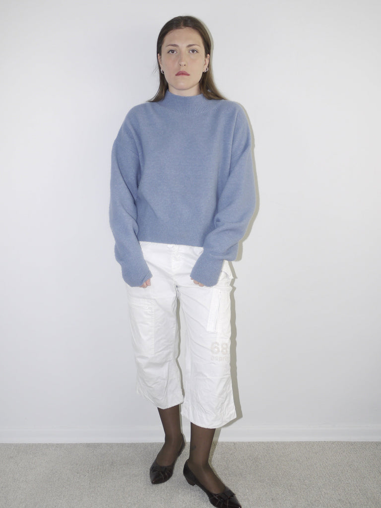 Model wearing a relaxed periwinkle blue knit sweater with structured shoulders, paired with white utility-style cropped pants — clean minimal outfit with soft color palette, shot against a neutral white background.