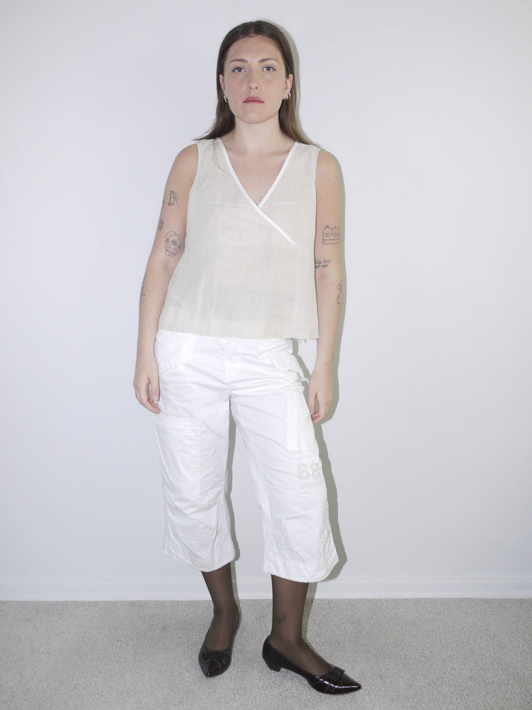 Model wearing sleeveless off-white linen wrap top with cropped white cargo pants, black tights and pointed ballet flats — minimal Y2K-inspired outfit, photographed against plain white background.