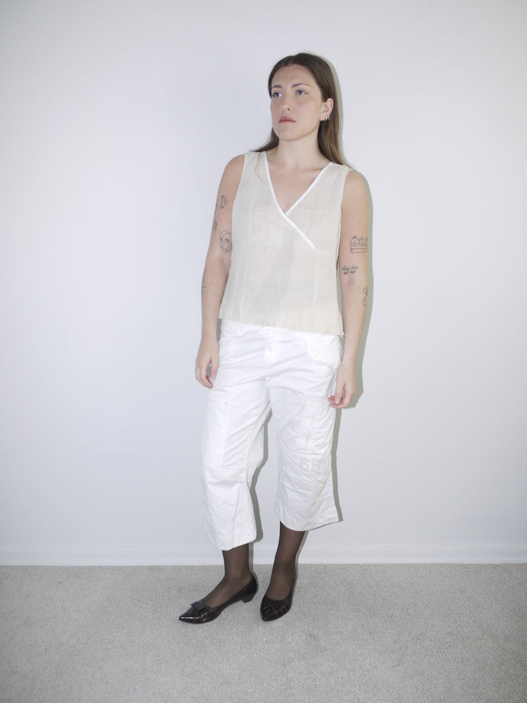 Model wearing sleeveless off-white linen wrap top with cropped white cargo pants, black tights and pointed ballet flats — minimal Y2K-inspired outfit, photographed against plain white background.