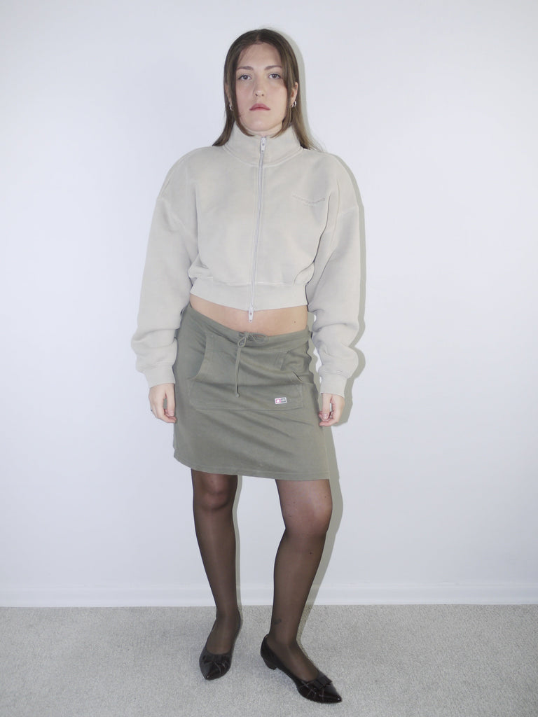 Vintage Y2K look featuring a cropped Alexander Wang zip-up fleece in beige, paired with an olive green mini skirt with front ruching and logo patch, styled with sheer tights and pointed flats.








