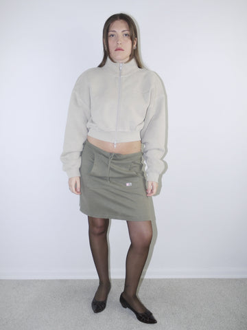 Vintage Y2K look featuring a cropped Alexander Wang zip-up fleece in beige, paired with an olive green mini skirt with front ruching and logo patch, styled with sheer tights and pointed flats.








